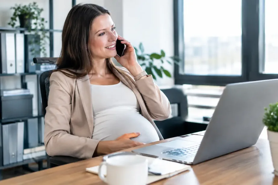 Kobieta w ciąży pracuje przy biurku w jasnym biurze i z uśmiechem rozmawia przez telefon