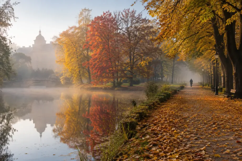Jesienny park ze stawem, w którym odbijają się złote i czerwone drzewa, oraz aleja pokryta liśćmi